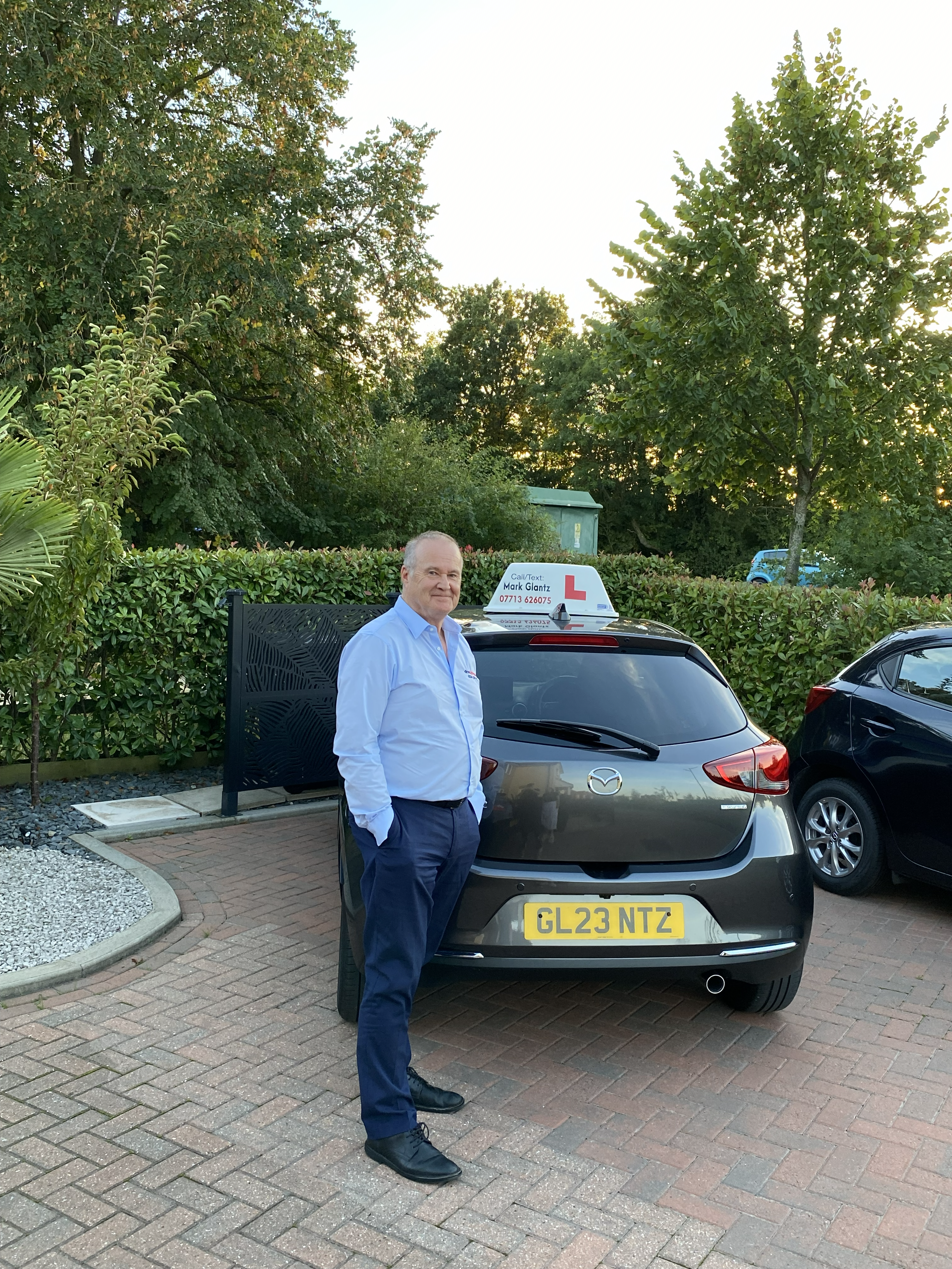 Smiling driving instructor stands beside a grey Mazda hatchback with a learner sign on top.